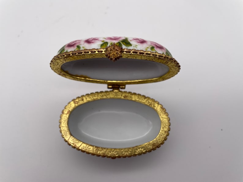 May include: Oval porcelain trinket box with a floral design and gold trim. The lid is open and the inside is white.
