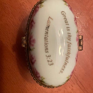 May include: A small, oval-shaped trinket box with a white porcelain exterior and gold trim. The box is decorated with pink roses and the text "Great is thy faithfulness. Lamentations 3:23."