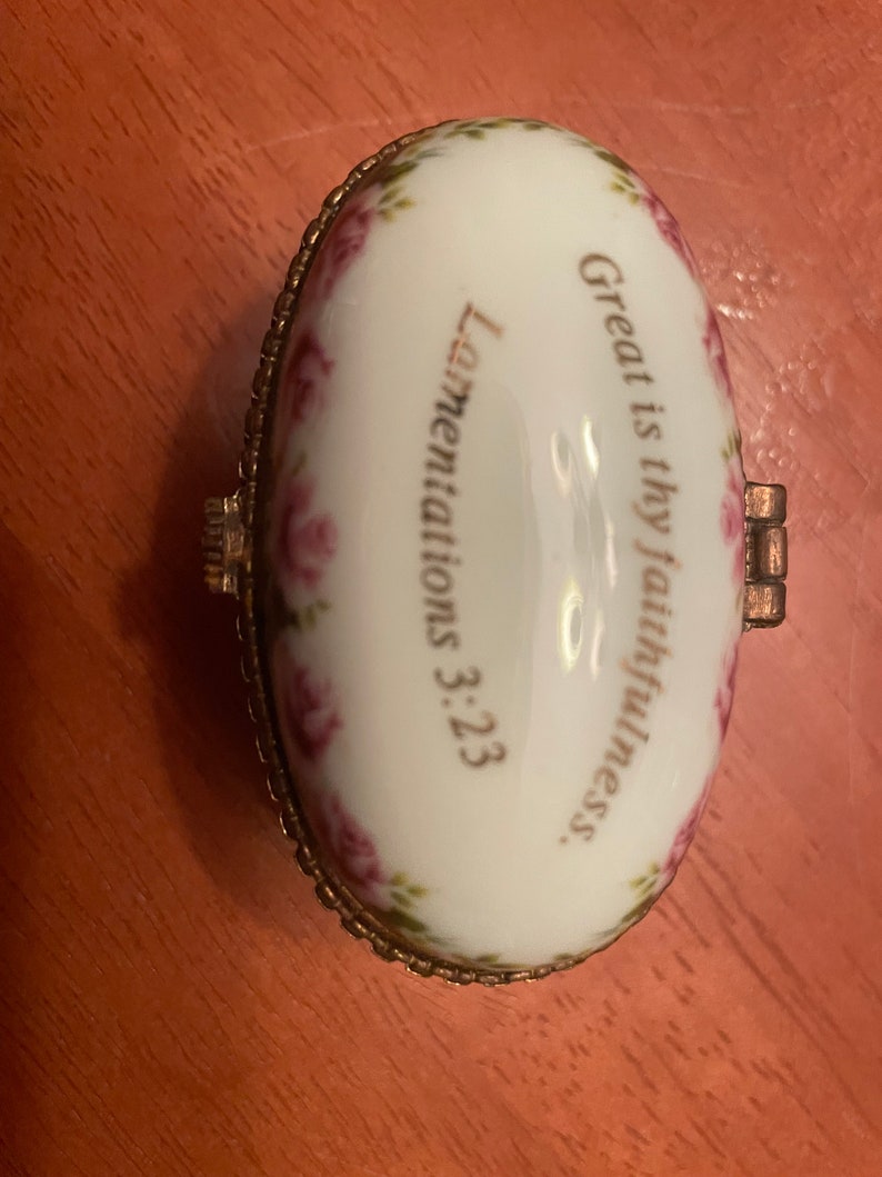 May include: A small, oval-shaped trinket box with a white porcelain exterior and gold trim. The box is decorated with pink roses and the text "Great is thy faithfulness. Lamentations 3:23."