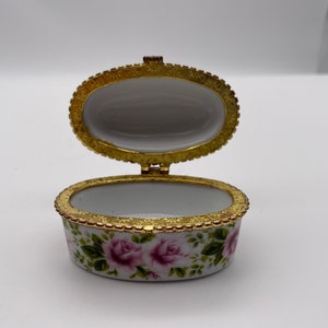 May include: A small oval trinket box with a floral design. The box is white with a gold trim and features pink roses and green leaves. The lid is open, revealing the empty interior.