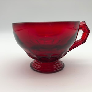 May include: A red glass cup with a footed base and a faceted handle. The cup is made of ruby red glass and has a vintage look.
