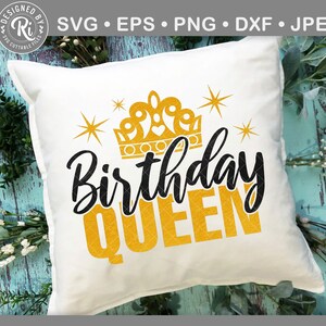 May include: A white square cushion featuring the text "Birthday QUEEN" in black and gold, with a gold crown and starbursts. The cushion is placed on a light blue wooden surface with foliage.
