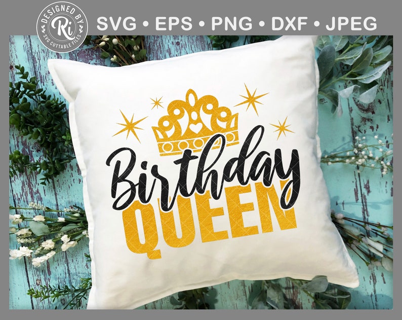May include: A white square cushion featuring the text "Birthday QUEEN" in black and gold, with a gold crown and starbursts. The cushion is placed on a light blue wooden surface with foliage.