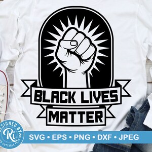 May include: White long-sleeve shirt with a black and white graphic of a raised fist inside a black arch with radiating lines. The words "BLACK LIVES MATTER" are printed below the fist in a banner style. White sneakers and blue jeans are in the background.