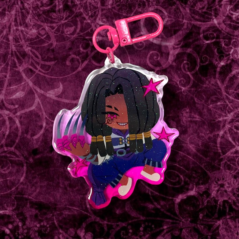 May include: A pink keychain featuring a cartoon character with dark hair, a star-shaped hair accessory, and a smiling expression. The character is wearing blue clothing with gold accents and holding pink flowers. The keychain has a pink clasp.