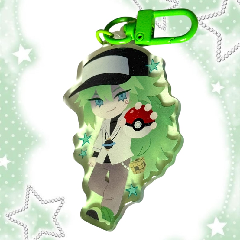 May include: A green and white anime-style keychain featuring a character holding a red and white Pok&eacute; Ball. The character has green hair, blue eyes, and is wearing a white jacket, black hat, and gray pants. The keychain has a green clasp.