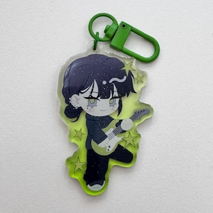 May include: A cartoon keychain featuring a character with dark hair and a black outfit playing a guitar. The character is set against a neon green background with star accents. The keychain has a green clasp.