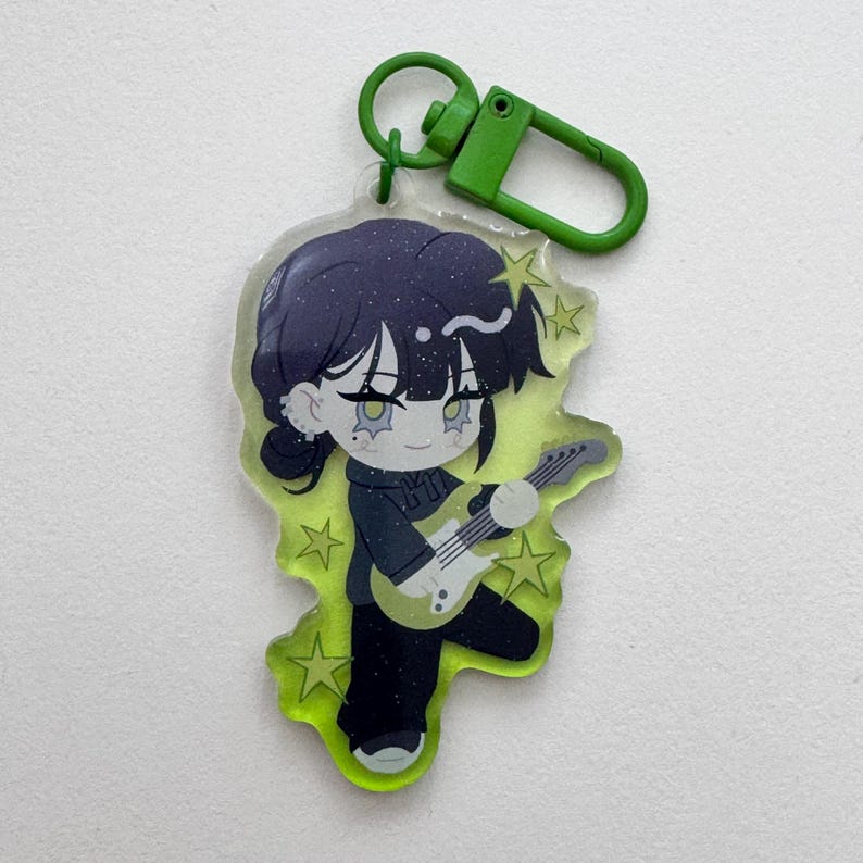 May include: A cartoon keychain featuring a character with dark hair and a black outfit playing a guitar. The character is set against a neon green background with star accents. The keychain has a green clasp.