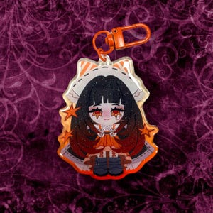 May include: A clear acrylic charm featuring a cartoon character with black hair, red eyes, and an orange dress. The charm has an orange clasp and star accents. The background is a dark purple with a swirling pattern.