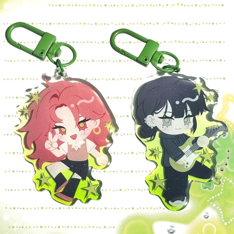 May include: Two acrylic keychains featuring cartoon characters. One keychain has a character with red hair, a peace sign, and gold earrings. The other keychain has a character with black hair playing a guitar. Both keychains have green clasps and star accents.