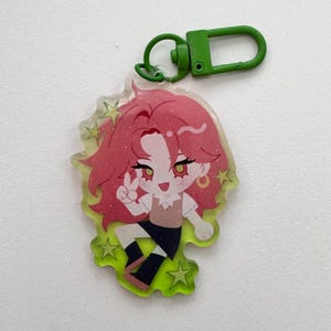 May include: A clear acrylic charm featuring a cartoon character with pink hair, gold earrings, and a peace sign. The charm has a green border with star accents and a green clasp.