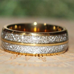 May include: A gold-toned ring featuring a central horizontal gold band flanked by two textured silver-colored sections. The outer edges of the ring are finished with smooth gold-toned borders, creating a layered metallic appearance.