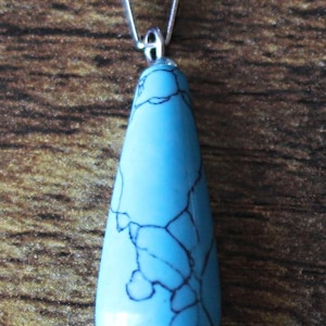 May include: A teardrop-shaped pendant made of turquoise with a silver bail. The turquoise is a light blue color with dark blue veins.