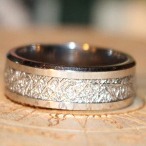 May include: A silver ring with a textured band and polished edges. The ring features a detailed, patterned design in the center, creating a striking contrast with the smooth borders. This piece of jewelry is suitable for everyday wear.