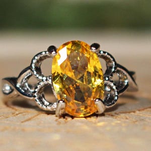 Könnte beinhalten: Ein silberfarbener Ring mit einem großen, oval geschliffenen, leuchtend gelben Edelstein, der von vier Krappen gehalten wird. Die Ringschiene ist mit einem komplizierten, durchbrochenen Metalldesign versehen, das sich seitlich erstreckt.
