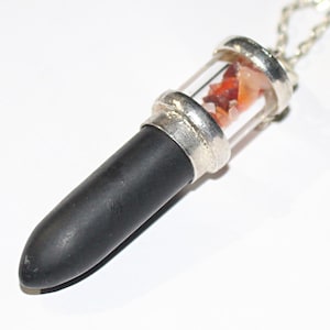 Può includere: Una collana con pendente con una fiala cilindrica di vetro riempita di pietre arancioni e rosse, sormontata da metallo argentato. La parte inferiore è un design liscio, nero, a forma di proiettile, sospeso da una catena d'argento.
