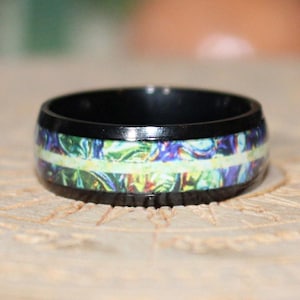 Puede incluir: Un anillo negro con una incrustación colorida y estampada. El anillo presenta una banda de tonos verdes, azules y morados, con una franja verde claro en el centro. El anillo está sobre un fondo neutro.