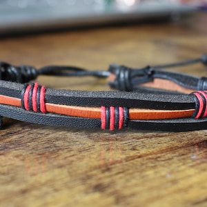 Może przedstawiać: Czarna skórzana bransoletka z cienkim pomarańczowym paskiem i czerwonymi przeszyciami. Bransoletka ma plecione skórzane zapięcie i jest regulowana. Stylowy dodatek do codziennego użytku.