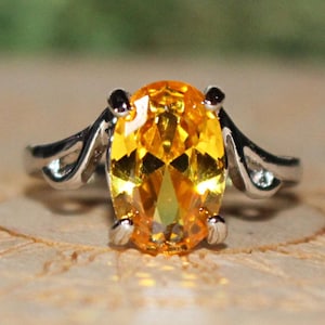 May include: A silver-toned ring featuring a large, oval-cut, vibrant yellow-orange gemstone held in a four-prong setting. The band displays a decorative, split-shoulder design that tapers towards the sides, creating a polished metallic finish.