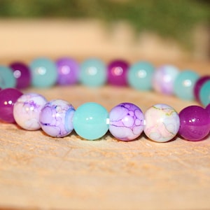 May include: A beaded bracelet featuring a variety of colorful beads. The bracelet includes beads in shades of purple, teal, and white with a marbled design. The bracelet is displayed on a wooden surface.