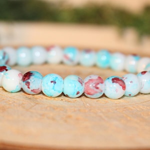May include: A beaded bracelet featuring round, light blue and white beads with red and blue accents. The bracelet is displayed on a wooden surface. Jewelry.