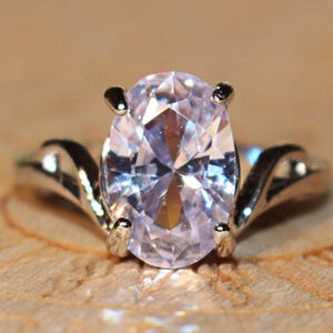 Puede incluir: Un anillo de tono plateado con una gran piedra preciosa de corte ovalado en color lavanda pálido, sujeta por cuatro garras. La banda del anillo tiene un acabado liso y pulido, curvándose hacia arriba para encontrarse con el engaste central.