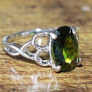 Könnte beinhalten: Ein silberner Ring mit einem ovalen, olivgrünen Edelstein. Der Ring hat ein aufwendiges, offenes Design am Band. Der Edelstein ist in einer Vier-Zacken-Fassung gefasst, auf einer Holzoberfläche.