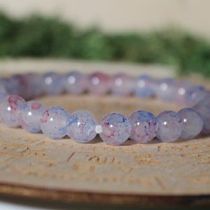 May include: A stretch bracelet composed of smooth, round, translucent beads. Each bead features a pale lavender base color with internal inclusions of vibrant red and blue speckles, creating a marbled effect throughout the circular jewelry piece.