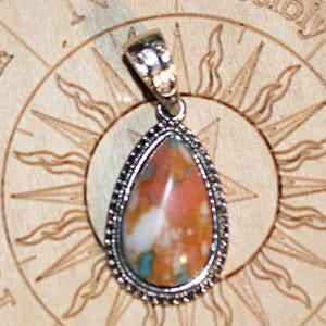 May include: A teardrop-shaped pendant featuring a polished multicolored stone with patches of orange, white, and teal. The stone is encased in a textured silver-toned metal setting with a decorative bail at the top for attachment to a chain.