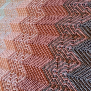 May include: A crocheted blanket with a gradient of colours from white to pink to brown. The blanket is made with a chevron pattern.