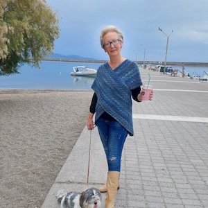 May include: A woman wearing a blue and grey patterned sweater, blue jeans, and brown boots walks a small white and grey dog on a leash. She is holding a pink cup with a green straw. The woman is walking on a paved path near a body of water.