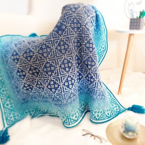 May include: A blue and white crocheted throw blanket with a geometric pattern. The blanket has a gradient of color, starting with a darker blue and fading to a lighter blue. The blanket is draped over a chair and has tassels on the ends.