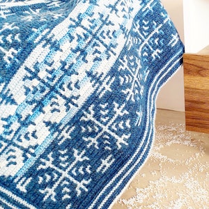 May include: A blue and white crocheted blanket with a geometric pattern. The blanket features a repeating design of stylized snowflakes and stripes in varying shades of blue and white. The blanket is folded over a surface.