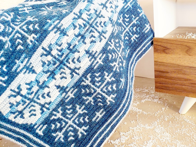May include: A blue and white crocheted blanket with a geometric pattern. The blanket features a repeating design of stylized snowflakes and stripes in varying shades of blue and white. The blanket is folded over a surface.