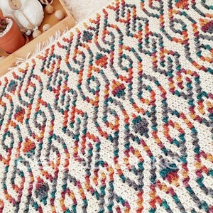 May include: A handmade, off-white crochet blanket with a repeating geometric pattern in teal, orange, gray, and burgundy. The blanket is displayed with yarn and craft supplies, suggesting a cozy, handcrafted theme.