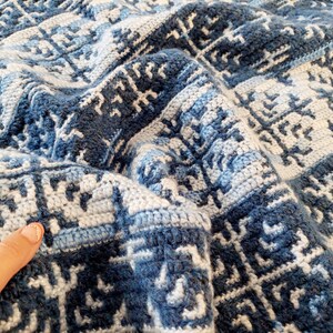 May include: A folded, handmade crochet blanket in shades of blue and white. The blanket features a geometric pattern with a textured appearance. The colour palette includes light and dark blues, and white, creating a visually appealing design.