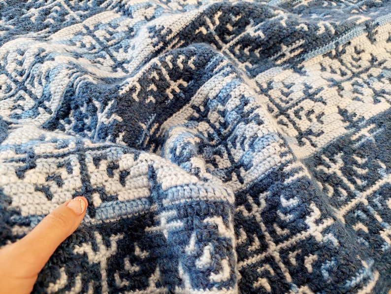May include: A folded, handmade crochet blanket in shades of blue and white. The blanket features a geometric pattern with a textured appearance. The colour palette includes light and dark blues, and white, creating a visually appealing design.