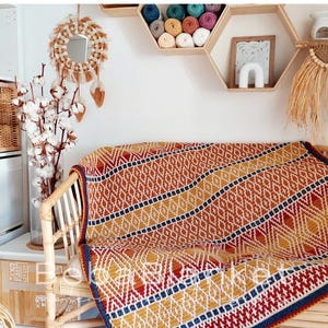 Può includere: Una coperta colorata con motivi geometrici drappeggiata su una sedia in rattan. La coperta presenta un motivo a rombi ripetuto nelle tonalità dell'arancione, del giallo, del rosso e del blu. Lo sfondo include mensole decorative, uno specchio e rami di cotone.