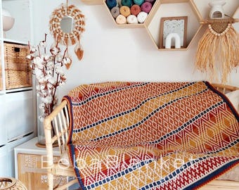 Puede incluir: Una colorida manta con estampado geométrico sobre una silla de ratán. La manta presenta un diseño de diamantes repetidos en tonos naranja, amarillo, rojo y azul. El fondo incluye estantes decorativos, un espejo y ramas de algodón.