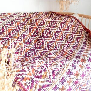 May include: A patterned blanket in shades of white, purple, orange, and brown, draped over a wooden sofa. The blanket's geometric design is complemented by a deep red border. The sofa has a natural wood frame. The blanket would be suitable for a double bed.