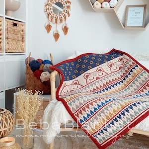 May include: A colorful crocheted blanket with a geometric pattern and a design of bears. The blanket is red, blue, yellow, and white. The blanket is draped over a white chair with a fluffy white throw.