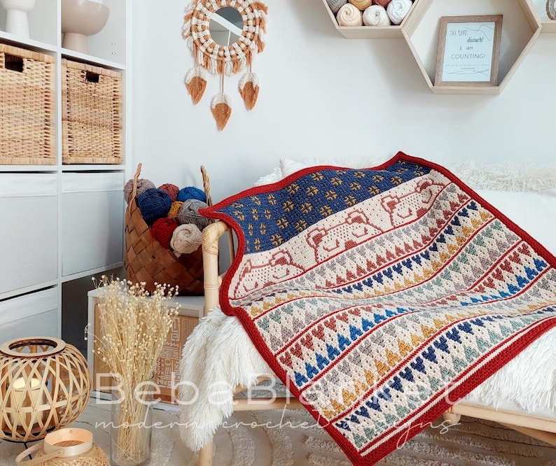May include: A colorful crocheted blanket with a geometric pattern and a design of bears. The blanket is red, blue, yellow, and white. The blanket is draped over a white chair with a fluffy white throw.