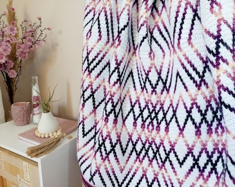 May include: A white crocheted blanket with a geometric pattern in shades of pink, purple, and black. The blanket has a gradient effect, with the pink and purple colors fading into each other.