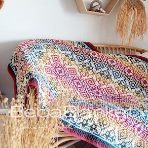 May include: A colorful, patterned blanket draped over a wicker chair. The blanket features a geometric design in shades of red, pink, yellow, blue, and white. The blanket's border is a dark red. The chair is made of light brown wicker.