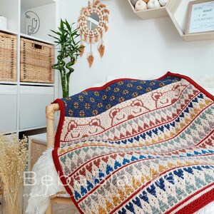 May include: A crocheted throw blanket with a geometric pattern in shades of red, blue, yellow, and white. The blanket features a repeating pattern of hearts and triangles. The blanket is draped over a chair with a white faux fur throw.