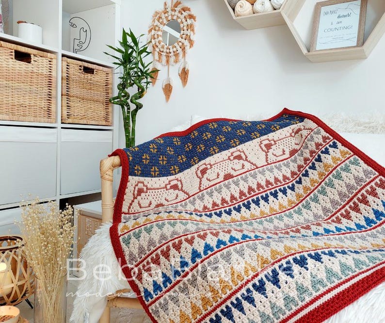 May include: A crocheted throw blanket with a geometric pattern in shades of red, blue, yellow, and white. The blanket features a repeating pattern of hearts and triangles. The blanket is draped over a chair with a white faux fur throw.