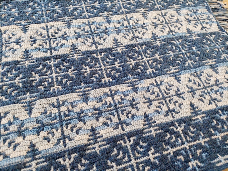 May include: A handmade, crocheted blanket in shades of blue and white. The blanket features a repeating geometric pattern with a fringe along one edge. The design incorporates squares and stylized tree shapes, creating a textured, cosy appearance.