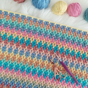 May include: A colorful crochet blanket with a variety of pastel colors, including pink, blue, yellow, and green. The blanket is made with a textured stitch pattern and is lying flat on a white surface. A pair of scissors and a crochet hook are also visible in the image.