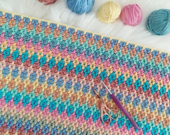 May include: A colorful crochet blanket with a variety of pastel colors, including pink, blue, yellow, and green. The blanket is made with a textured stitch pattern and is lying flat on a white surface. A pair of scissors and a crochet hook are also visible in the image.