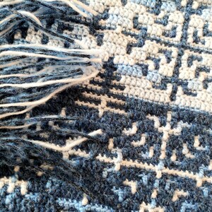 May include: Close-up of a handmade crochet blanket with a geometric pattern in shades of blue and white. The blanket has a fringe of white yarn along one edge. The design features a repeating square motif.